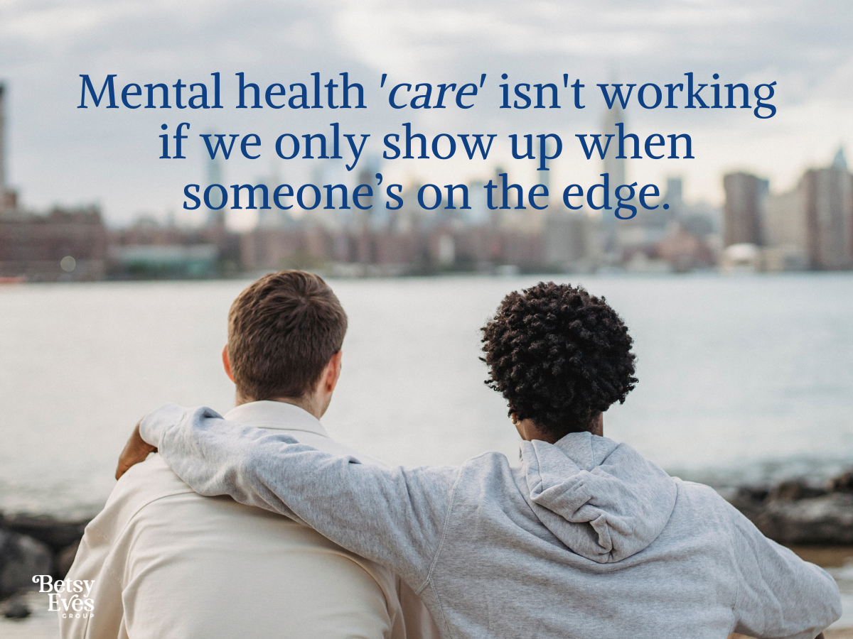 Image of two people sitting on a bench looking out at a city across water.  One person has their arm around the other.  Text overlay that reads: Mental health 'care' isn’t working if we only show up when someone’s on the edge.